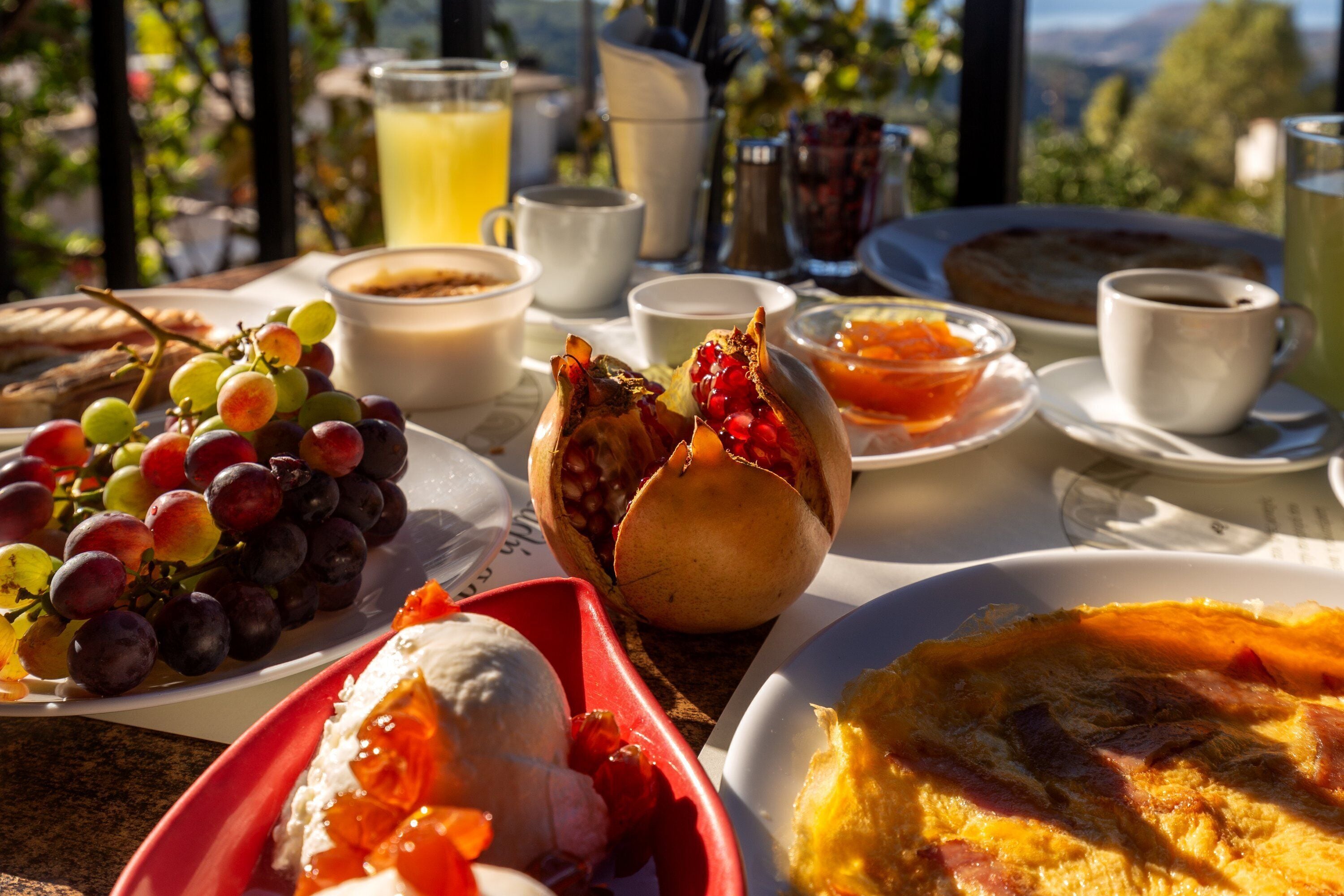 A breakfast table with various food items and drinks, including grapes, pomegranate, and a quiche, with a scenic view in the background.