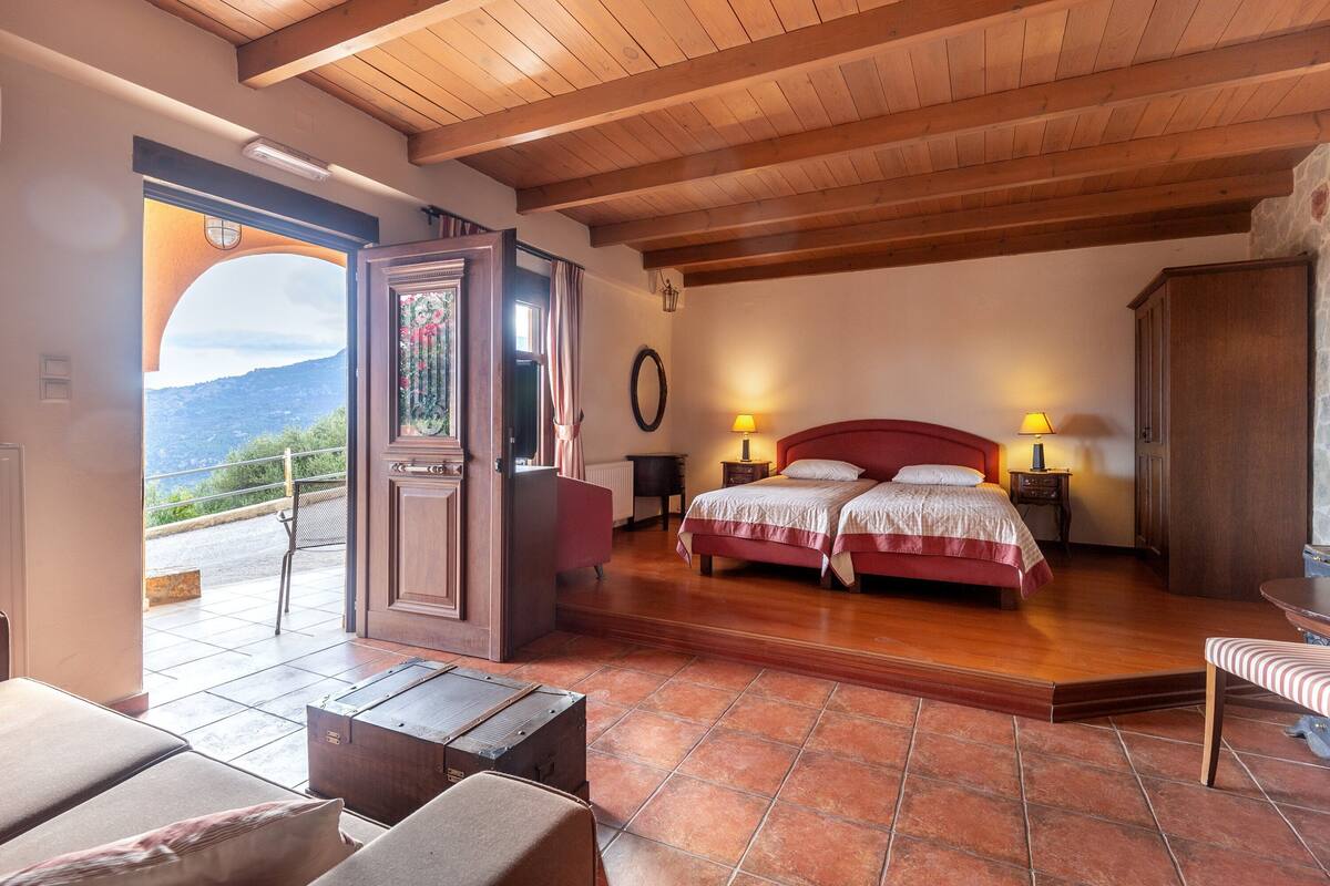 A bedroom with a wooden ceiling, a double bed, two bedside tables with lamps, a view of the outdoors, and a small seating area with a bench.