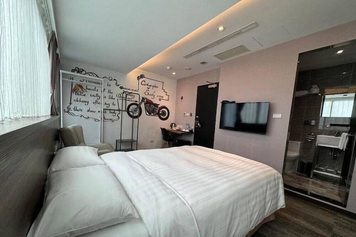 Una habitación de hotel moderna con una cama grande, un escritorio con silla, un televisor montado y un box de ducha de vidrio.