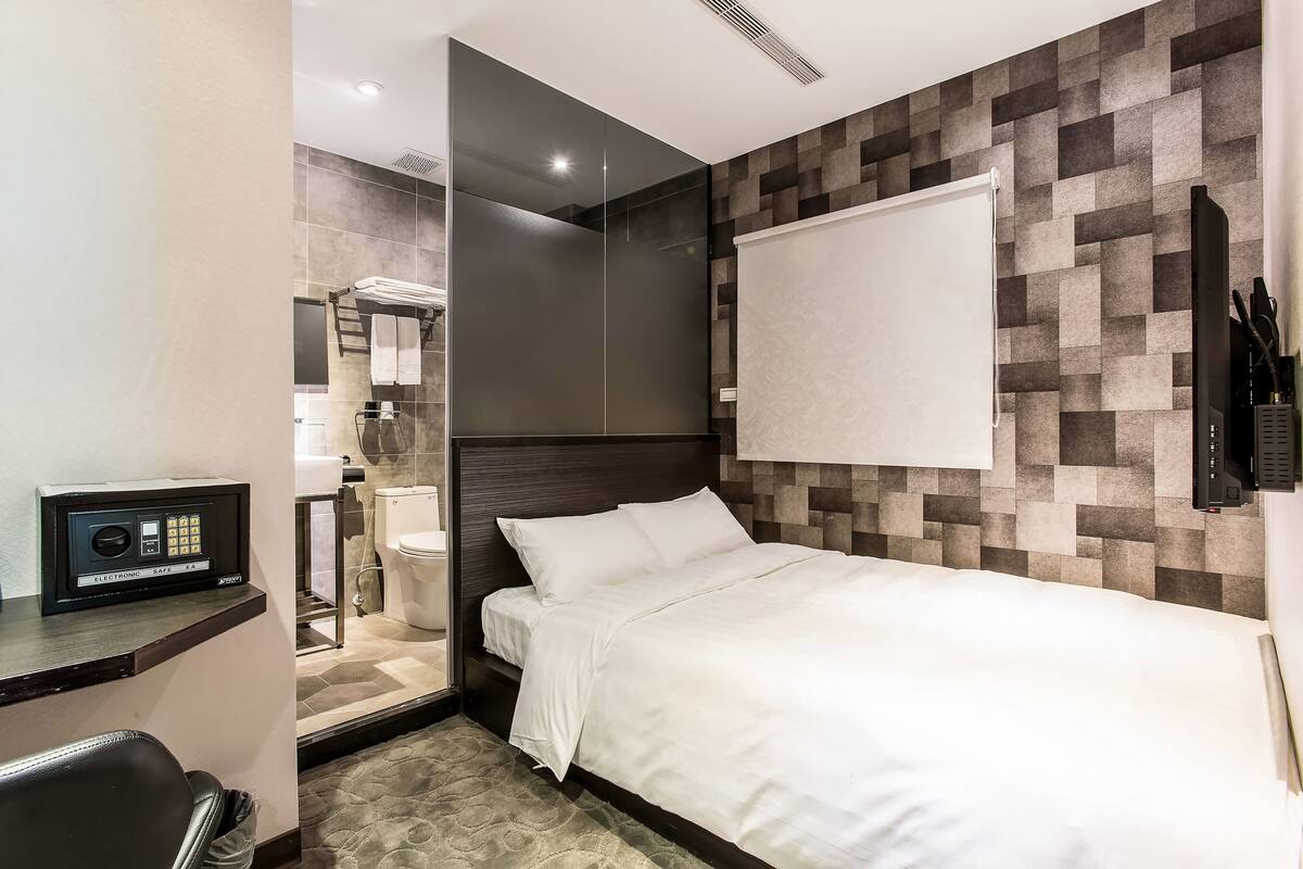 Habitación de hotel con cama, escritorio, espejo, zona de ducha y televisor montado en la pared.