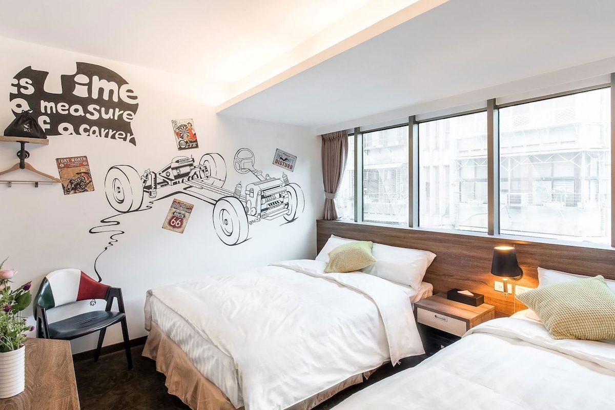 Una habitación de hotel con dos camas, un mural en la pared, una ventana con cortinas, una mesita de noche y una silla.