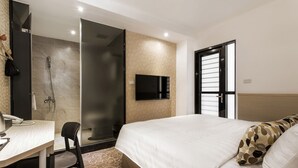 Quarto de hotel com cama, escrivaninha, cadeira, área de chuveiro e televisão.