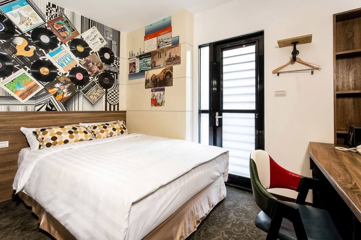 Habitación de hotel con cama, escritorio, silla, puerta y pared decorada con diversos objetos.