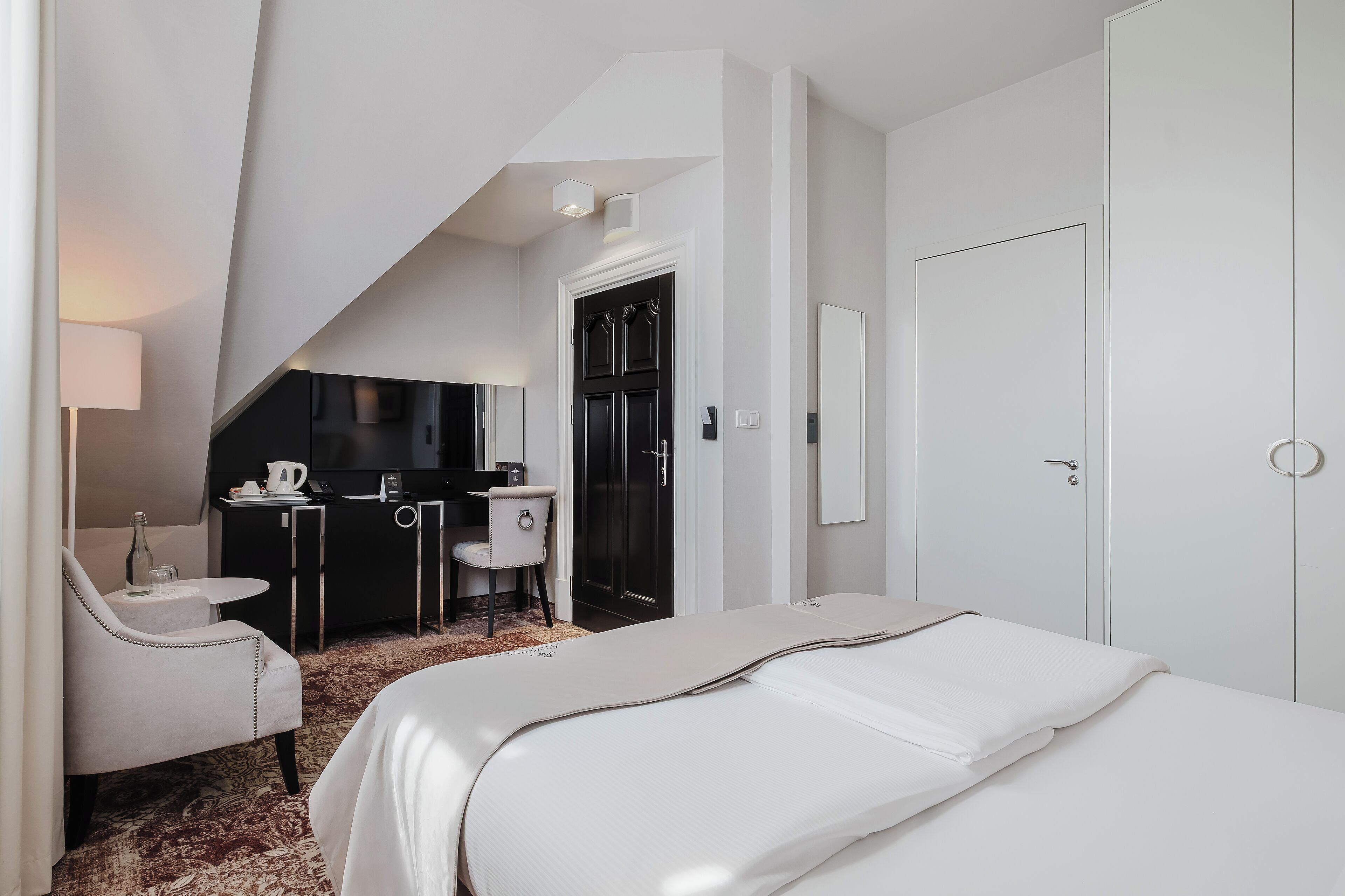 A hotel room with a bed, a desk with a chair, a small table with a bottle, and a TV.