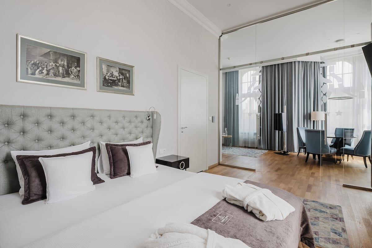 A hotel room with a large bed, a tufted headboard, two framed pictures on the wall, a desk with a chair, and a view through a glass door to a dining area.