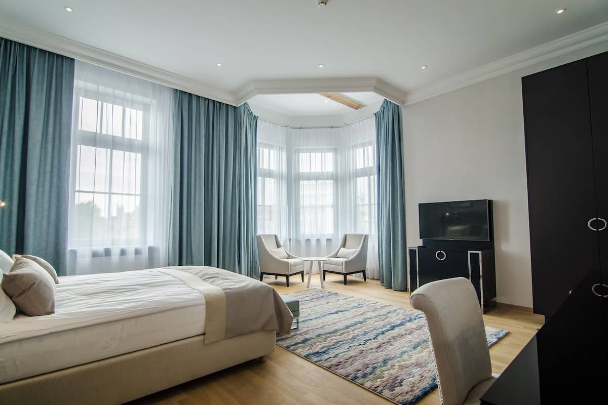 A modern hotel room with a large bed, two armchairs, a TV, and a bay window with curtains.
