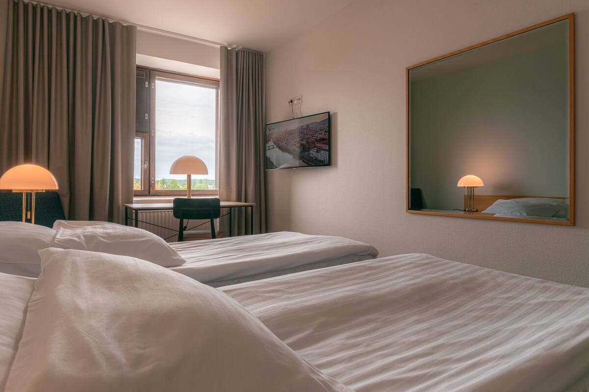 A hotel room with two beds, a desk, a chair, a large mirror, and a window with curtains.