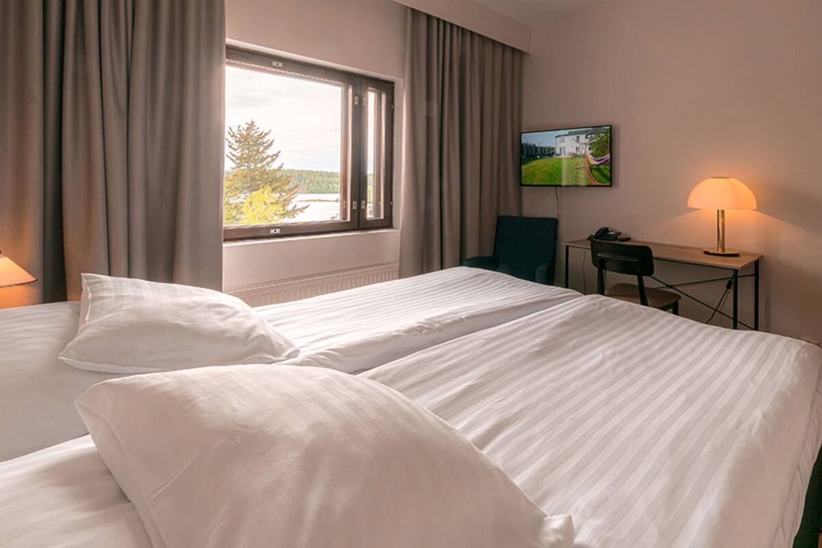 A hotel room with two beds, a desk, a lamp, a TV, and a window with curtains.