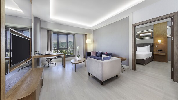 Ein modernes Hotelzimmer mit großem Flachbildfernseher, einem Schreibtisch mit Stuhl, einer Couch, einem Bett und einem Balkon mit Bergblick.