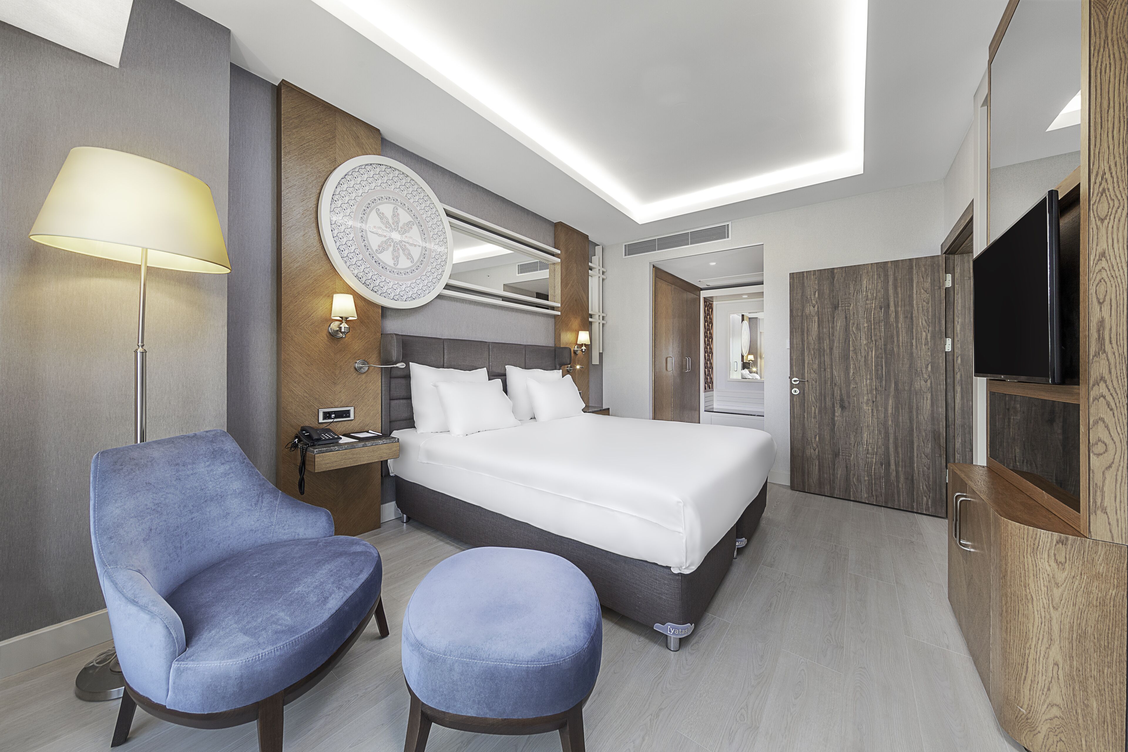 Una habitación de hotel moderna con cama, escritorio, silla, televisor y un espejo grande.