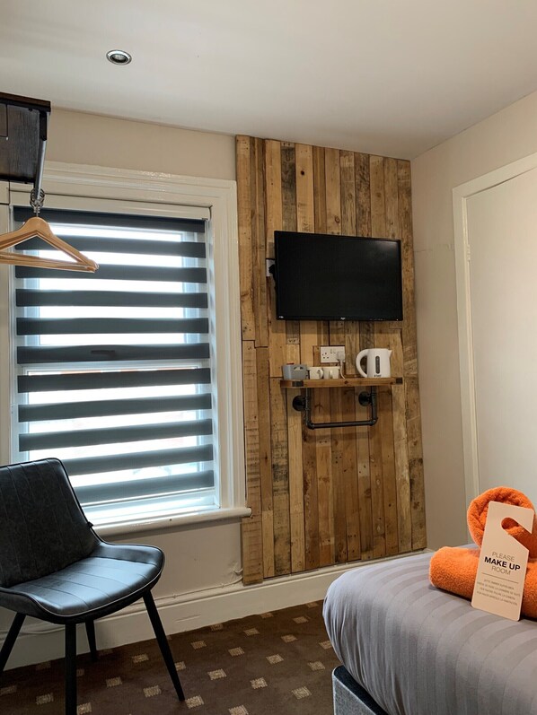 Une chambre d’hôtel avec un panneau mural en bois, un téléviseur à écran plat fixé au-dessus d’une étagère sur laquelle se trouve une machine à café, une chaise et un lit agrémenté d’un oreiller orange.
