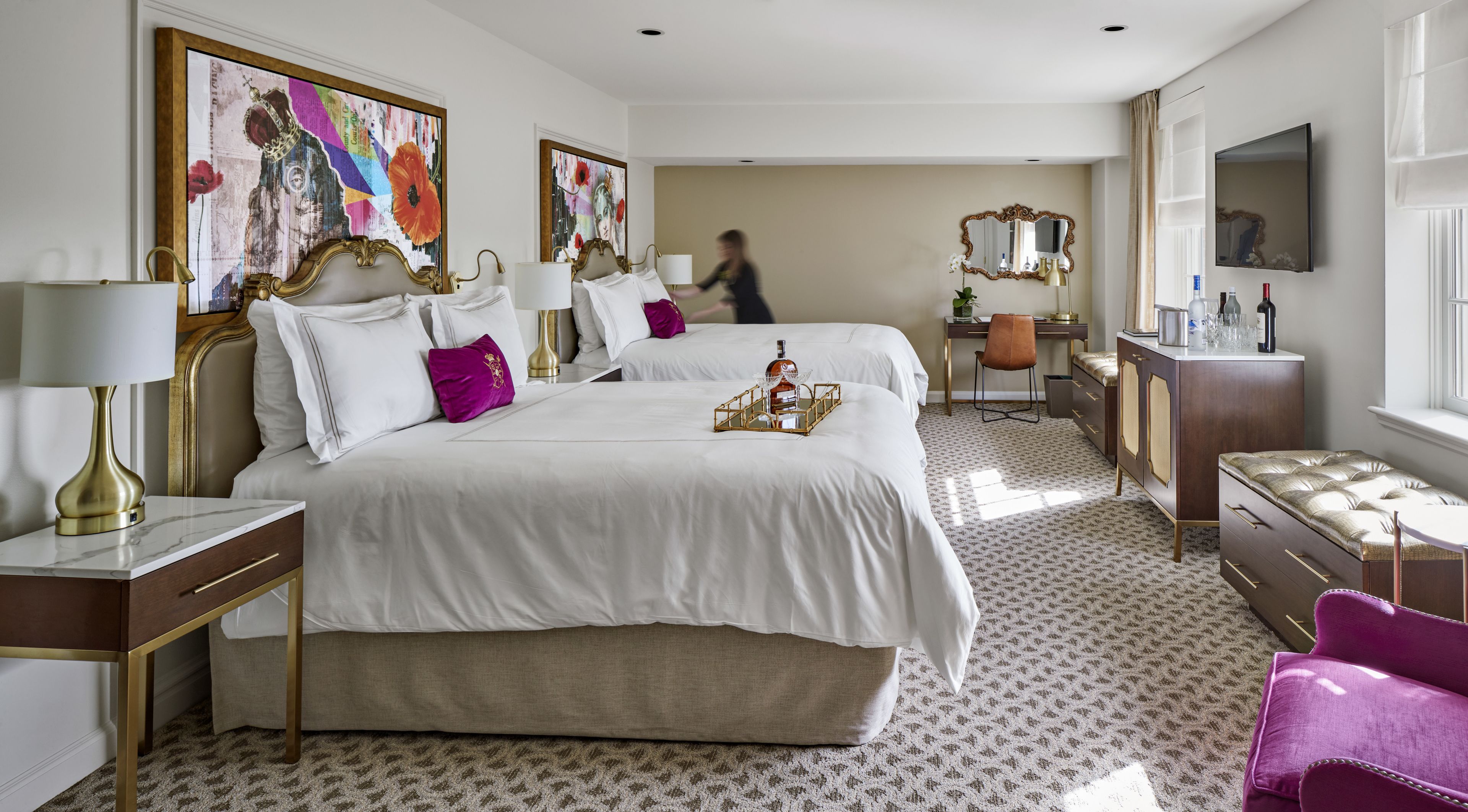 A hotel room with two beds, a desk, a chair, a TV, and artwork on the walls.