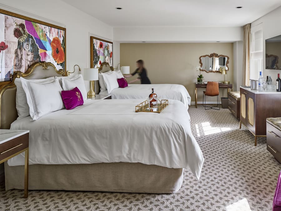 A hotel room with two beds, a desk, a chair, a TV, and artwork on the walls.