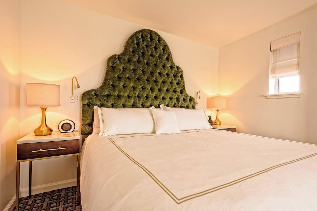A neatly made bed with a tufted headboard, bedside tables with lamps, and a window with blinds.