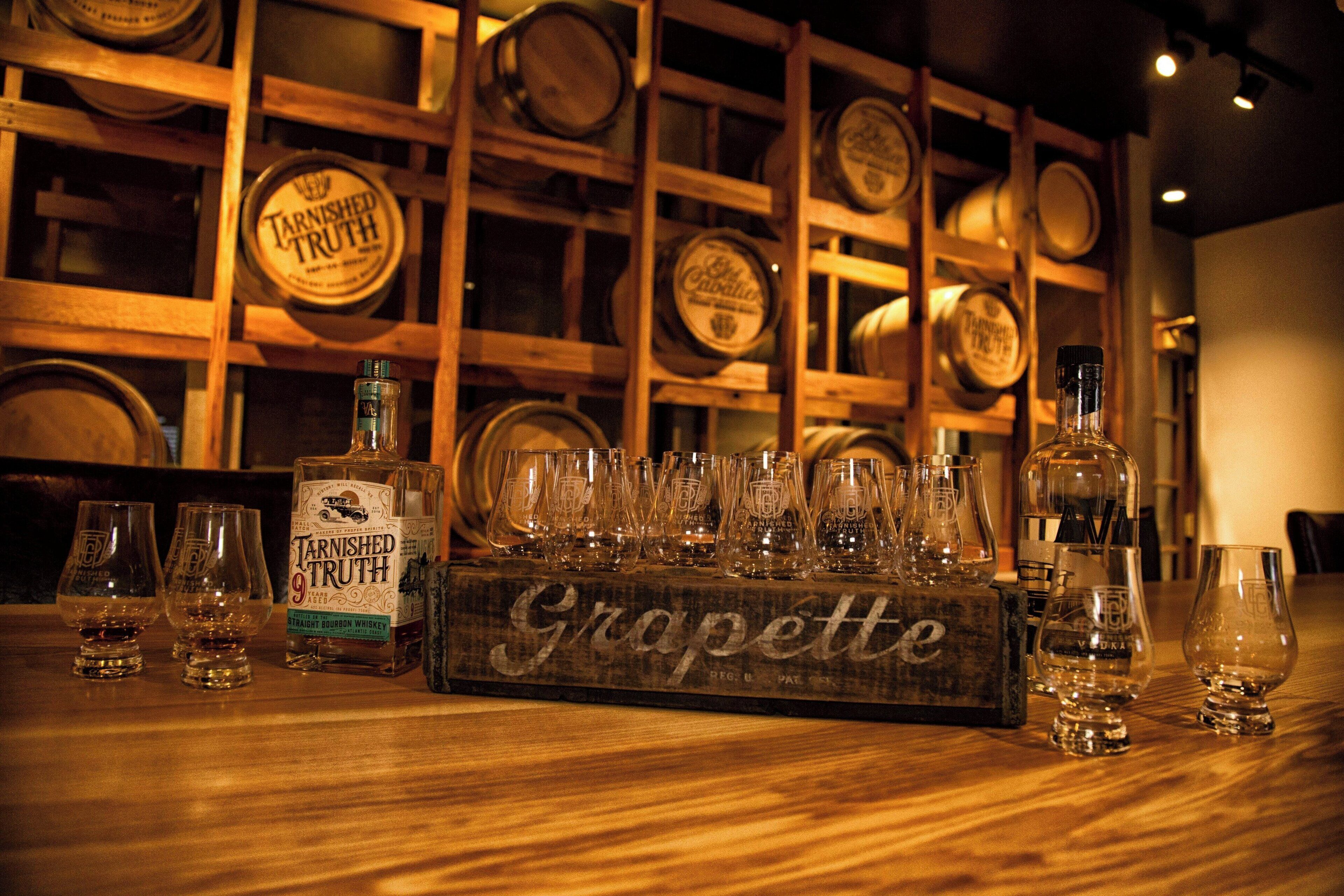 Una barra de madera con una botella de Tannished Truth y una caja de madera con la etiqueta 'Grapette', junto a varios vasos.