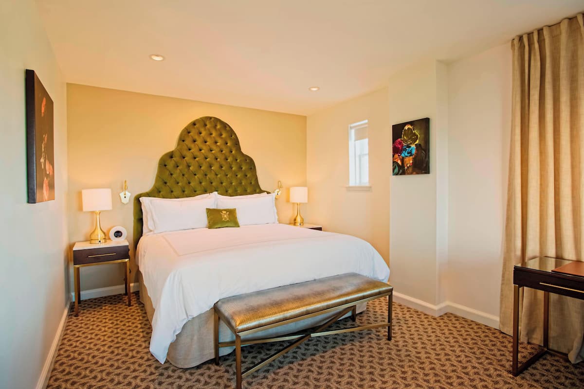 A hotel room with a tufted headboard, a large bed with white linens, two bedside tables with lamps, a bench, patterned carpet, and a window with curtains.