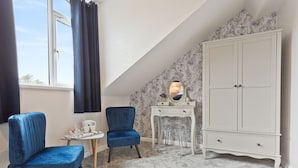 Une pièce avec un plafond en pente, une armoire blanche, deux chaises bleues, une petite table avec un miroir et un papier peint à motifs.