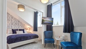 Une chambre à coucher avec un lit, deux chaises, une petite table, un téléviseur et une fenêtre avec des rideaux.