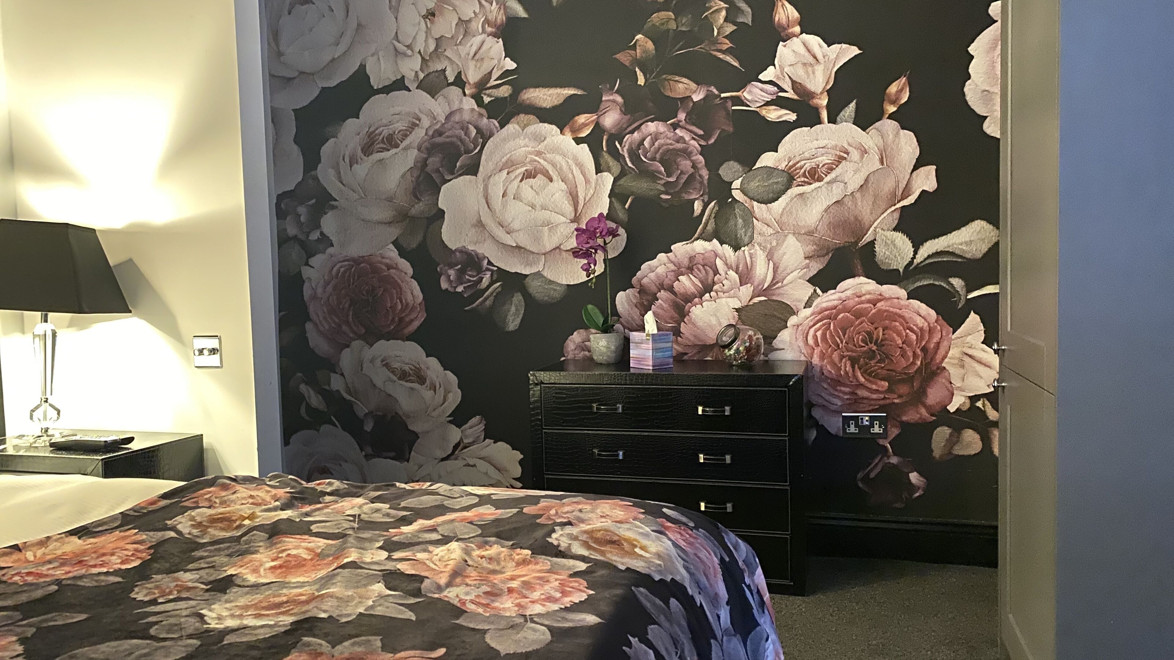 Ein Schlafzimmer mit blumigem Wandpapier, einem Bett mit Blumendecke, einem Schminktisch und einer Lampe.