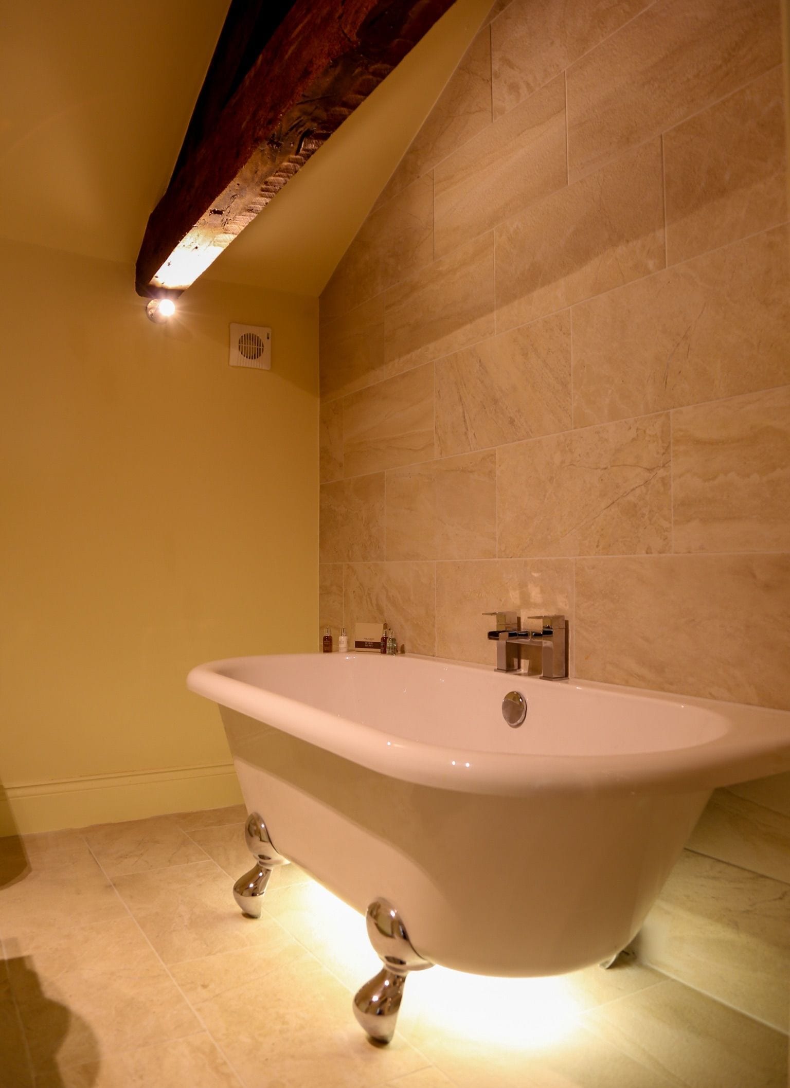 A bathroom with a freestanding bathtub, tiled walls, and a wooden ceiling beam.