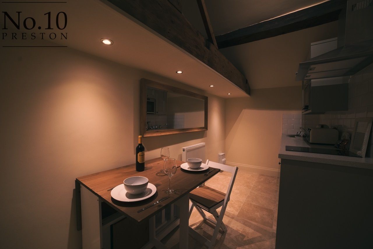 A compact kitchen with a wooden table set for two, a wine bottle, and a bowl. A white chair is placed at the table. The kitchen has a modern design with a large window and a built-in oven.