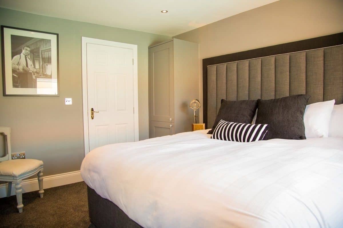 A neatly arranged bedroom with a large bed, a bedside table with a lamp, a framed picture on the wall, and a door.