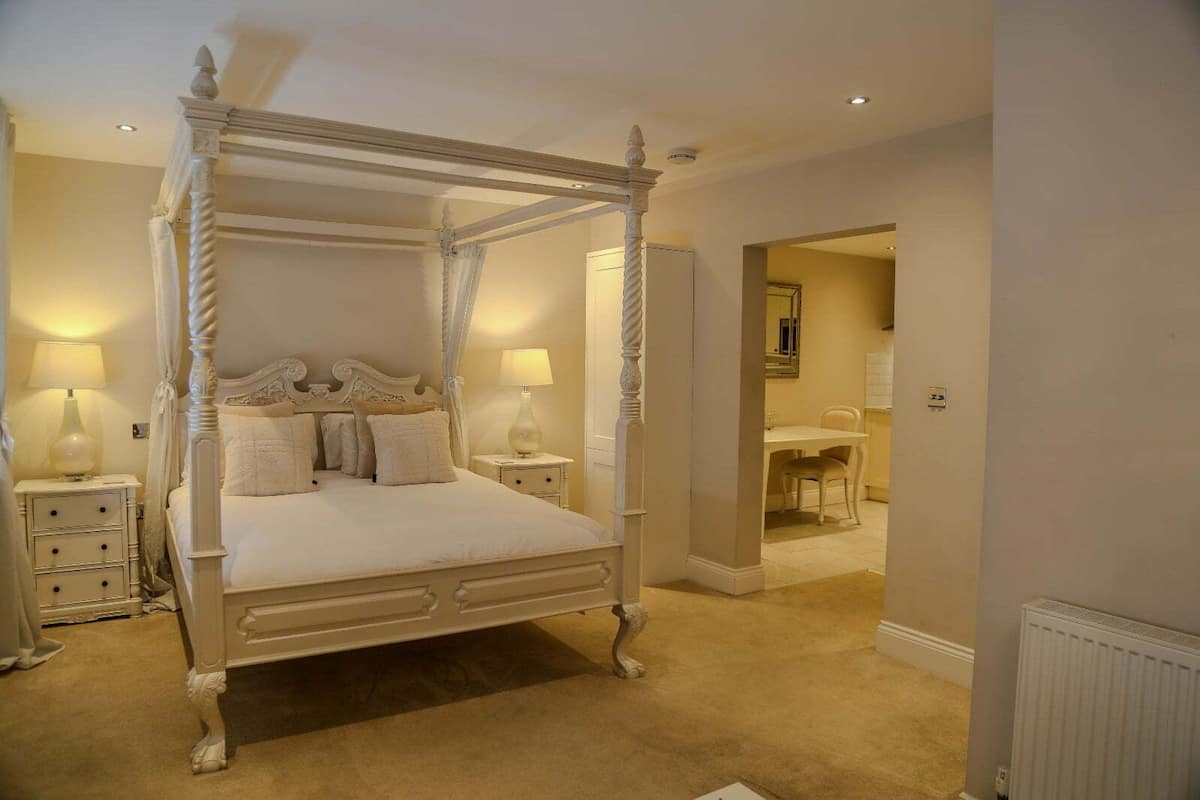 A four-poster canopy bed with a classic design, a bedside table with a lamp, and a chair in the adjacent room.