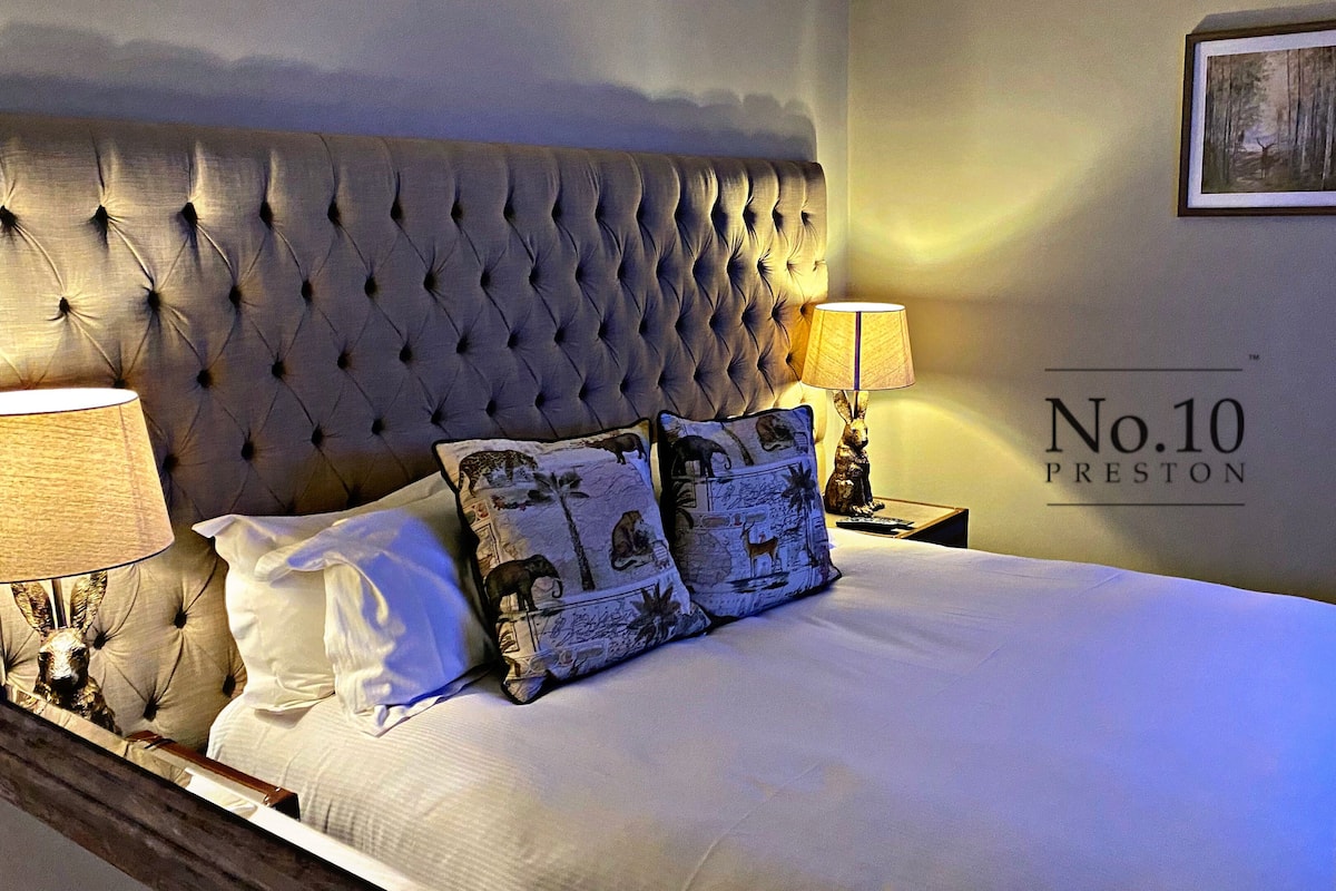 A hotel room with a tufted headboard, two pillows, a lamp, and a wall sign reading "No.10 Preston".