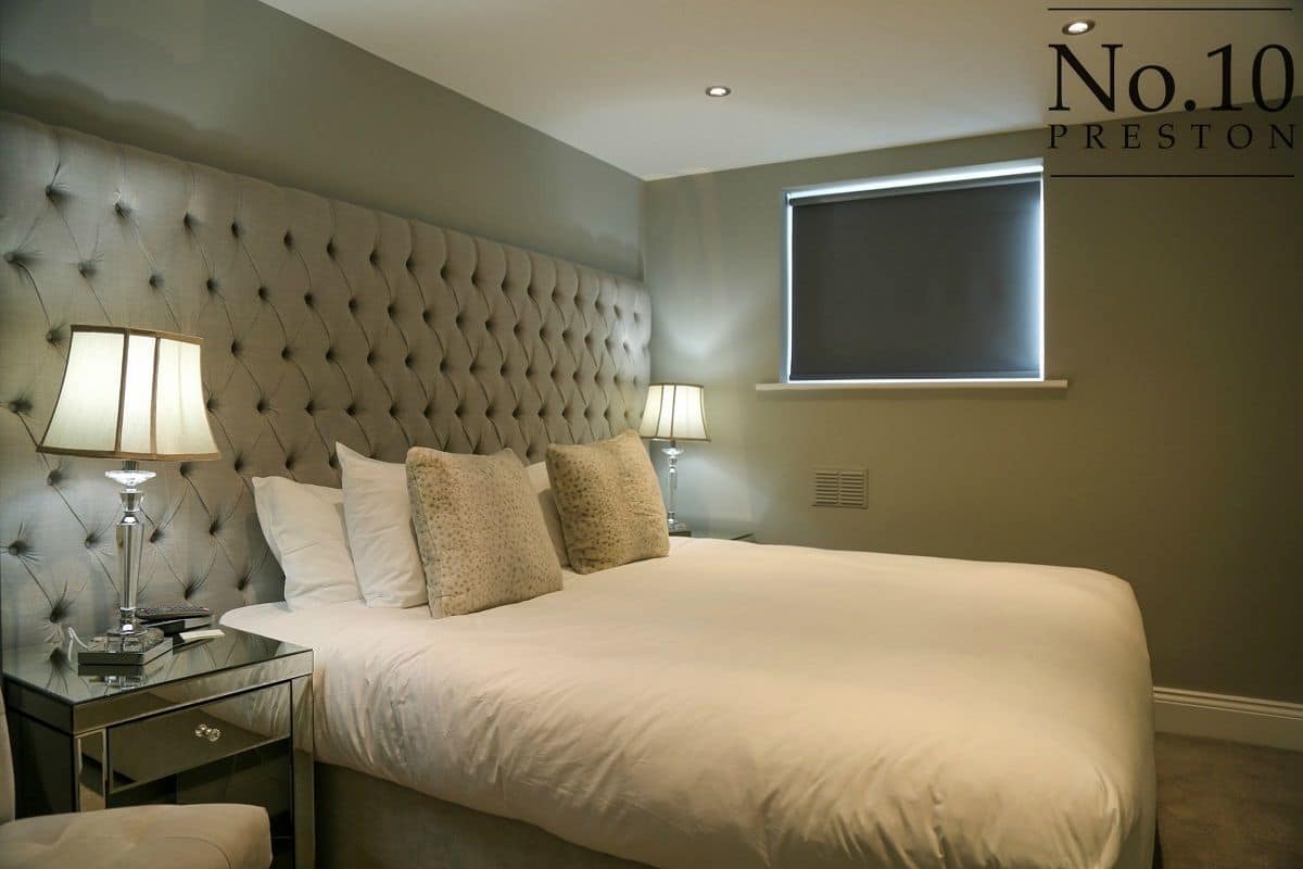 A bedroom with a tufted headboard, a large bed, two bedside lamps, and a window with blinds.