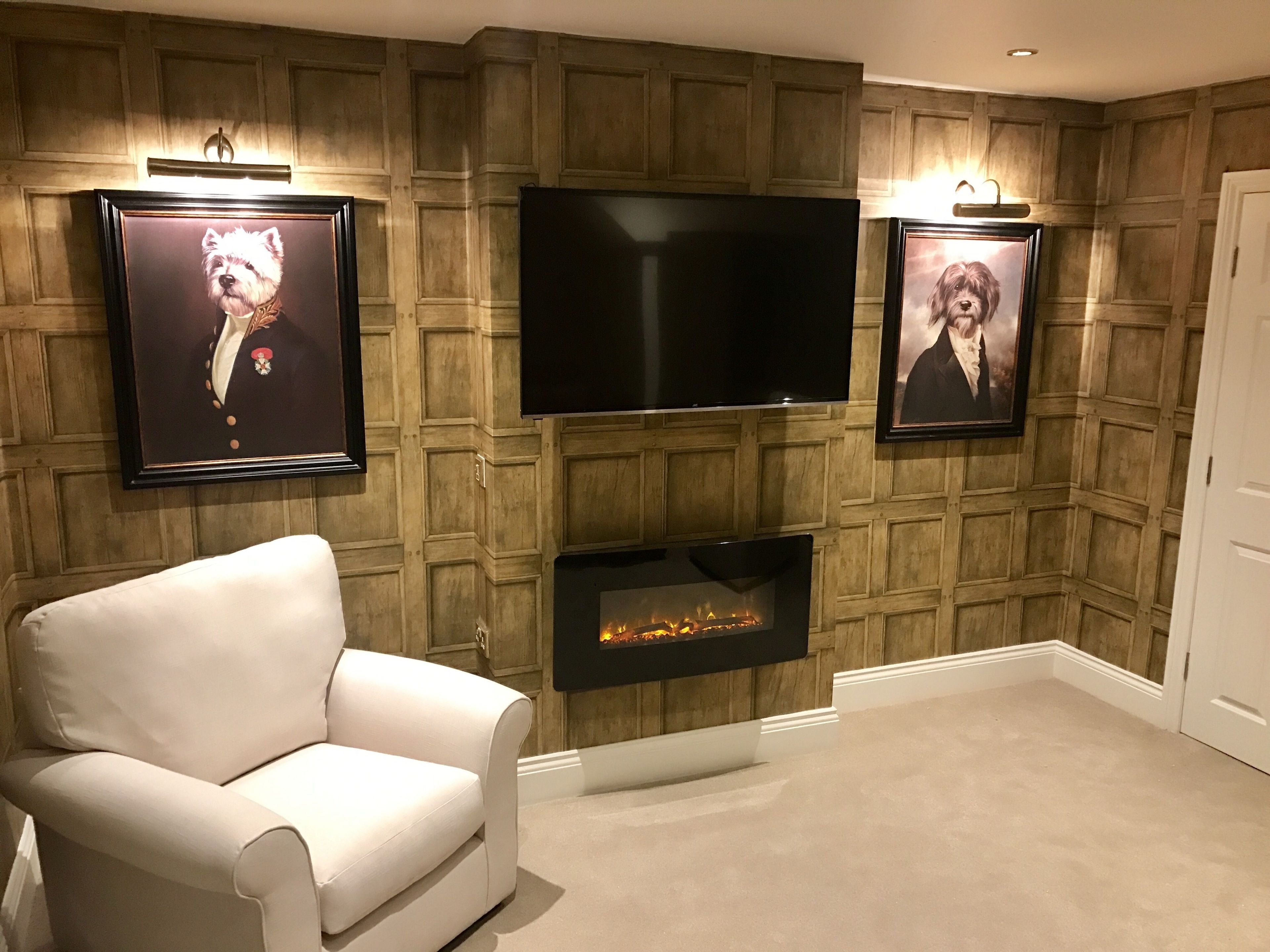 A living room with a flat-screen TV mounted on a wooden paneled wall, flanked by framed pictures. A white leather armchair is positioned in front of a fireplace.