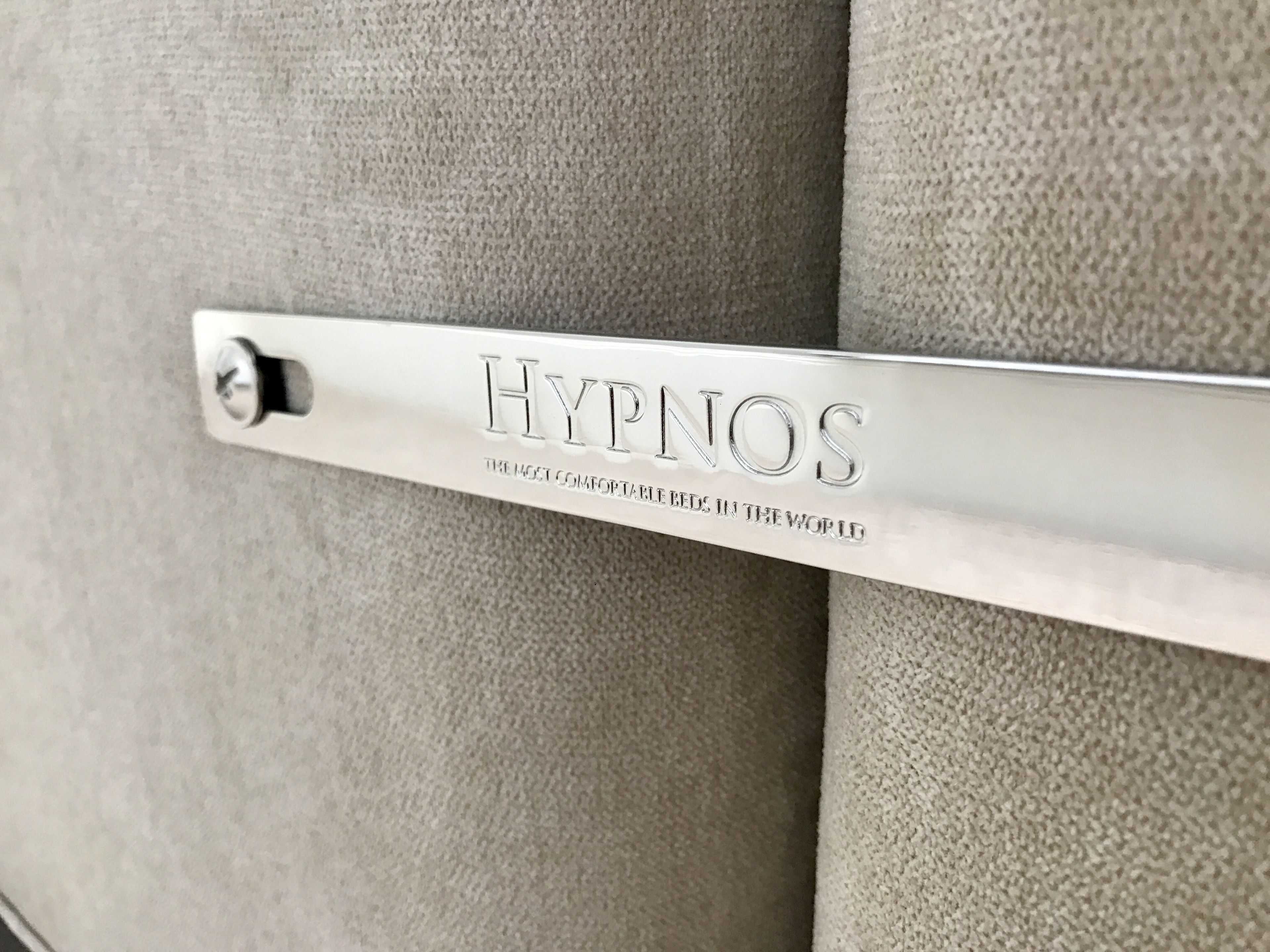 Une plaque métallique portant le mot « HYPNOS » et la phrase « LES LITS LES PLUS CONFORTABLES DU MONDE », le tout sur un fond en tissu texturé.