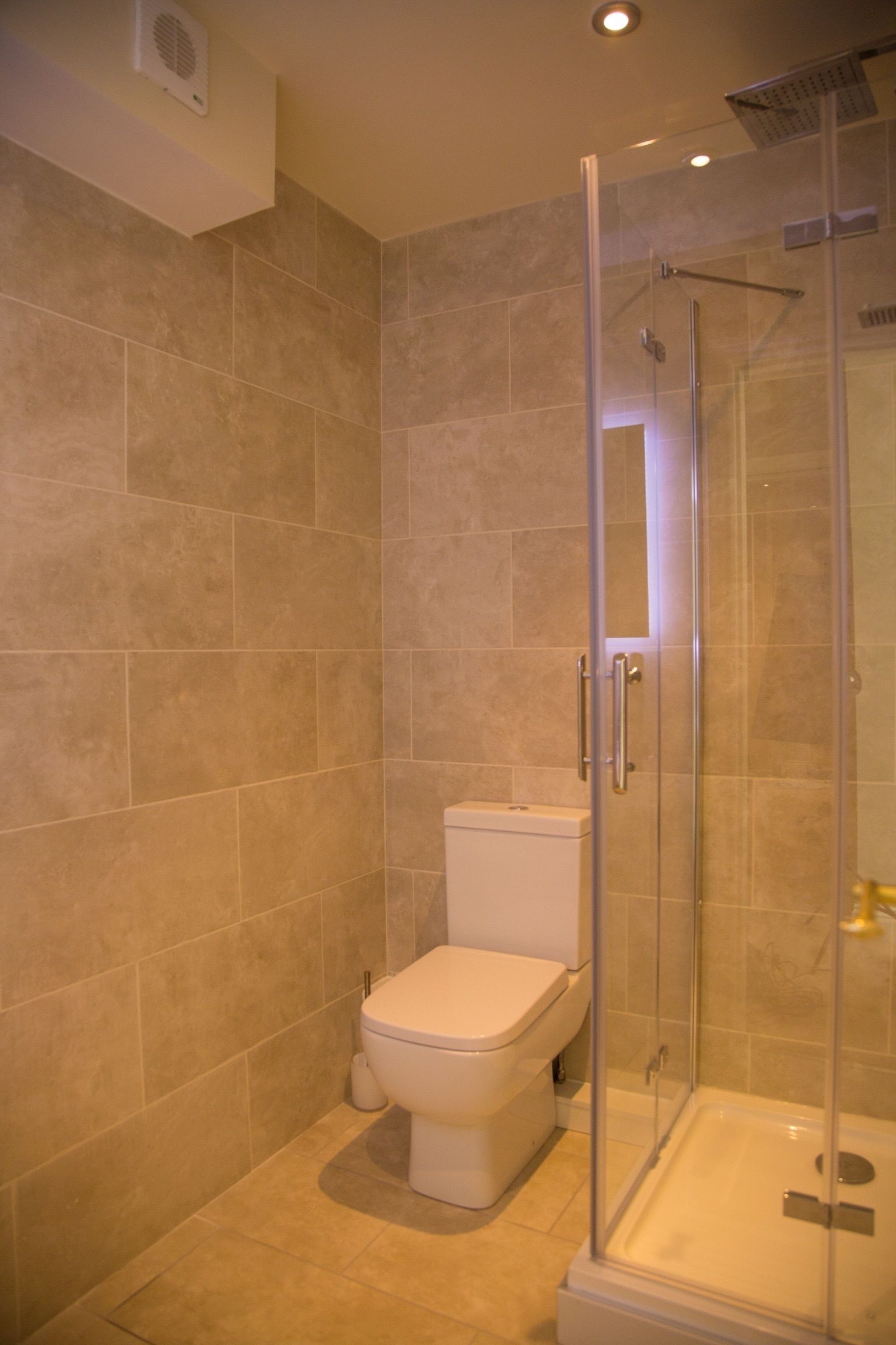 Un baño con azulejos color beige, un inodoro y una zona de ducha con mampara de vidrio.
