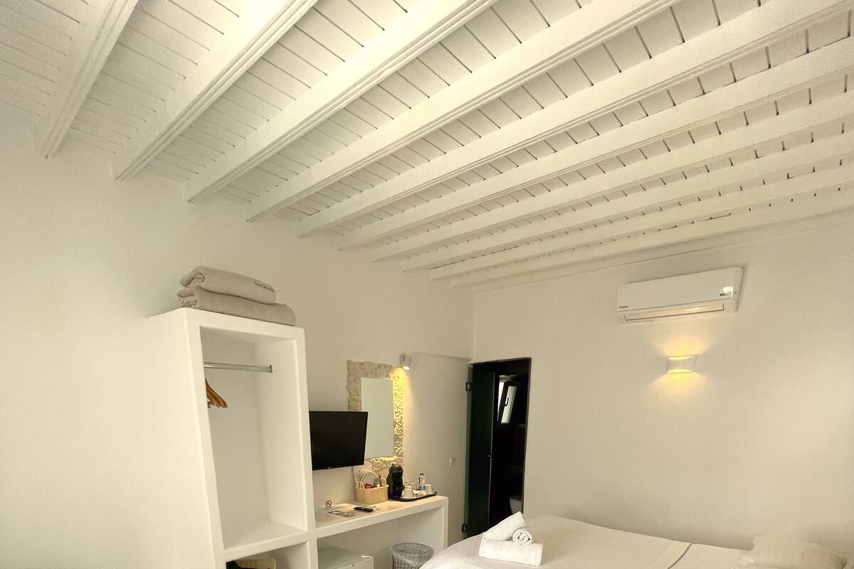 Quarto de hotel com uma cama, uma escrivaninha, um guarda-roupa e uma televisão.