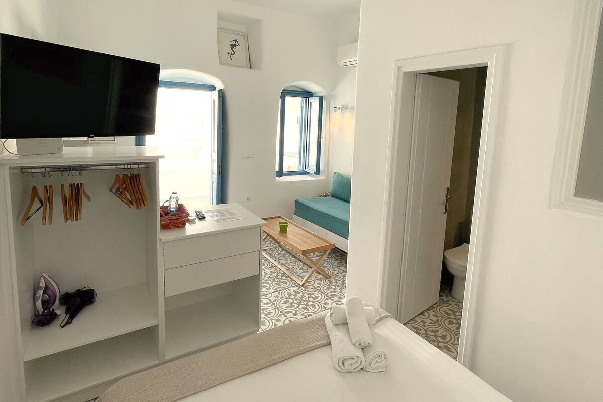 Quarto de hotel com cama, televisão, escrivaninha com cadeira, armário e banheiro visível por uma porta aberta.