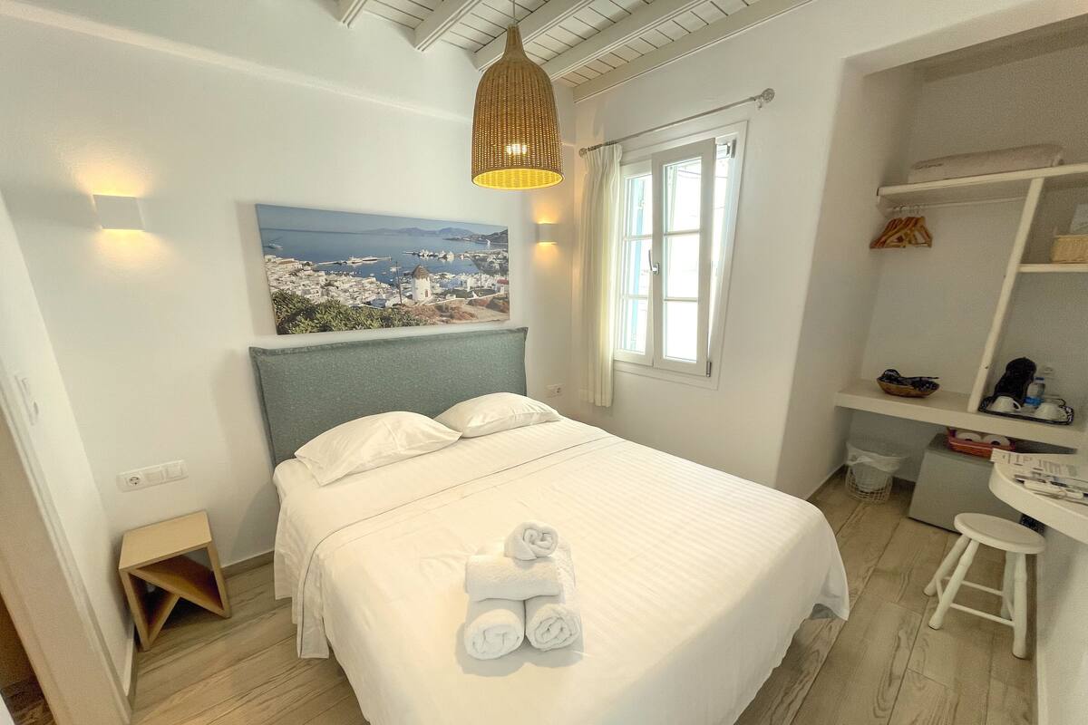 Quarto bem organizado com uma cama, escrivaninha e uma cadeira. Há uma janela com cortinas, um lustre e um quadro na parede.