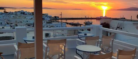 A rooftop terrace with white furniture and a view of a sunset over a coastal town.