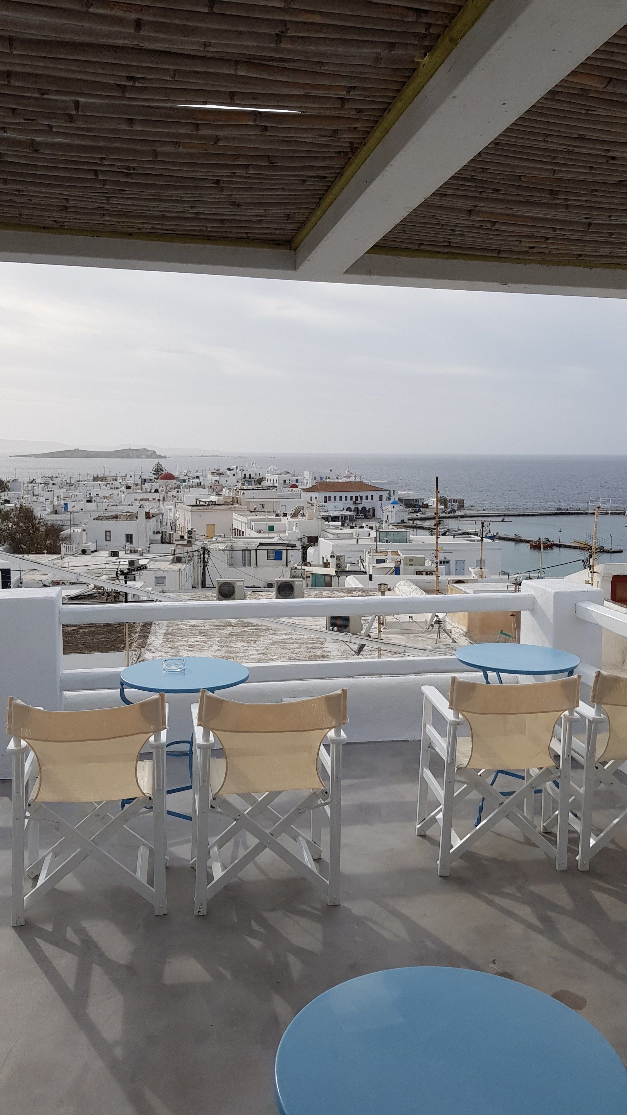 Une terrasse dotée de chaises blanches et de tables bleues, offrant une vue sur un paysage urbain côtier avec ses bâtiments et la mer.