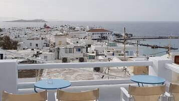Une terrasse dotée de chaises blanches et de tables bleues, offrant une vue sur un paysage urbain côtier avec ses bâtiments et la mer.