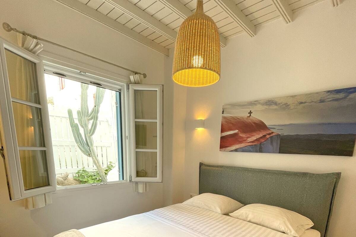 Uma cama bem arrumada com toalhas dobradas, uma janela grande com vista para um cacto e um quadro de uma paisagem na parede.