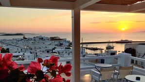 A terrace with a view of a coastal town at sunset, featuring white buildings, a marina, and red flowers. - Hotel Lefteris (Mykonos)