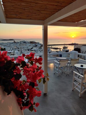 A terrace with a view of a coastal town at sunset, featuring white buildings, a marina, and red flowers. - Hotel Lefteris (Mykonos)