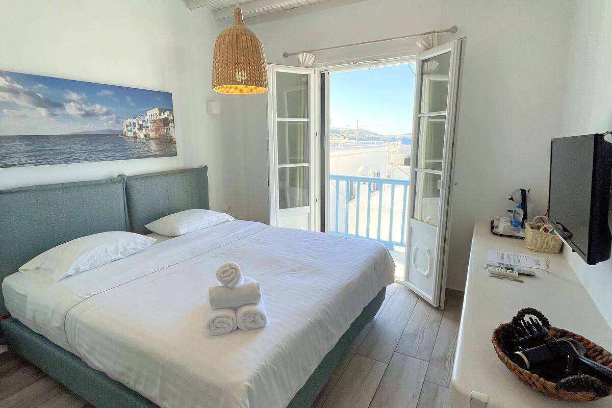 Quarto de hotel com cama grande, varanda com vista, escrivaninha com televisão e um quadro na parede.