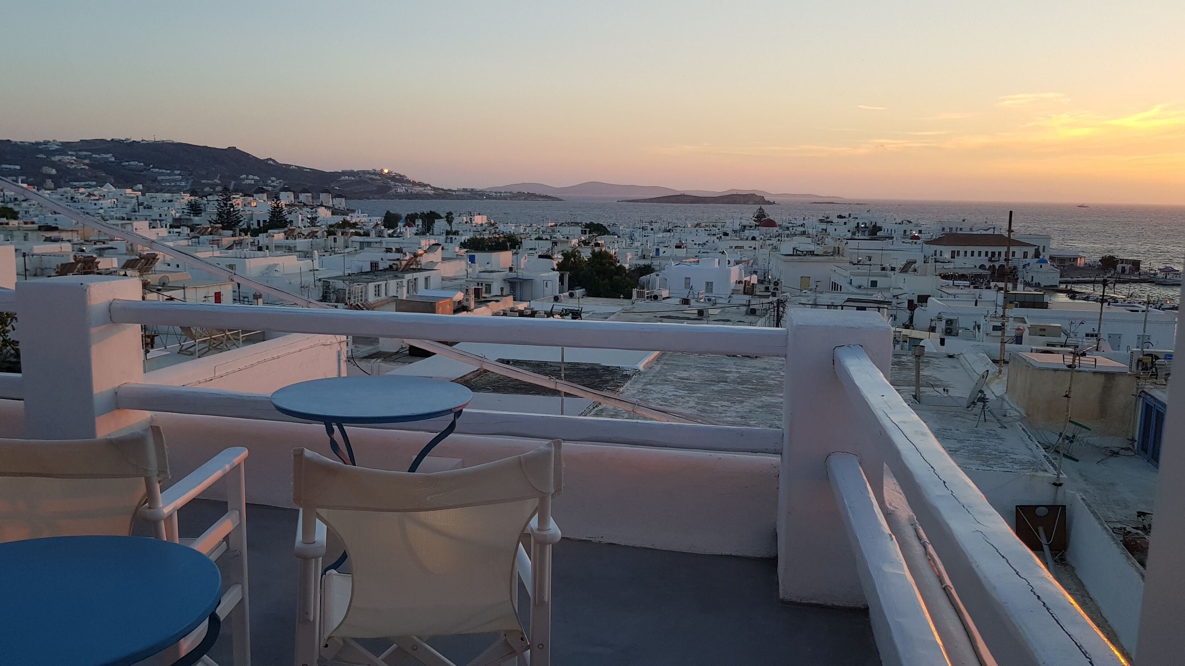 A balcony with a table and chairs overlooking a coastal town at sunset.