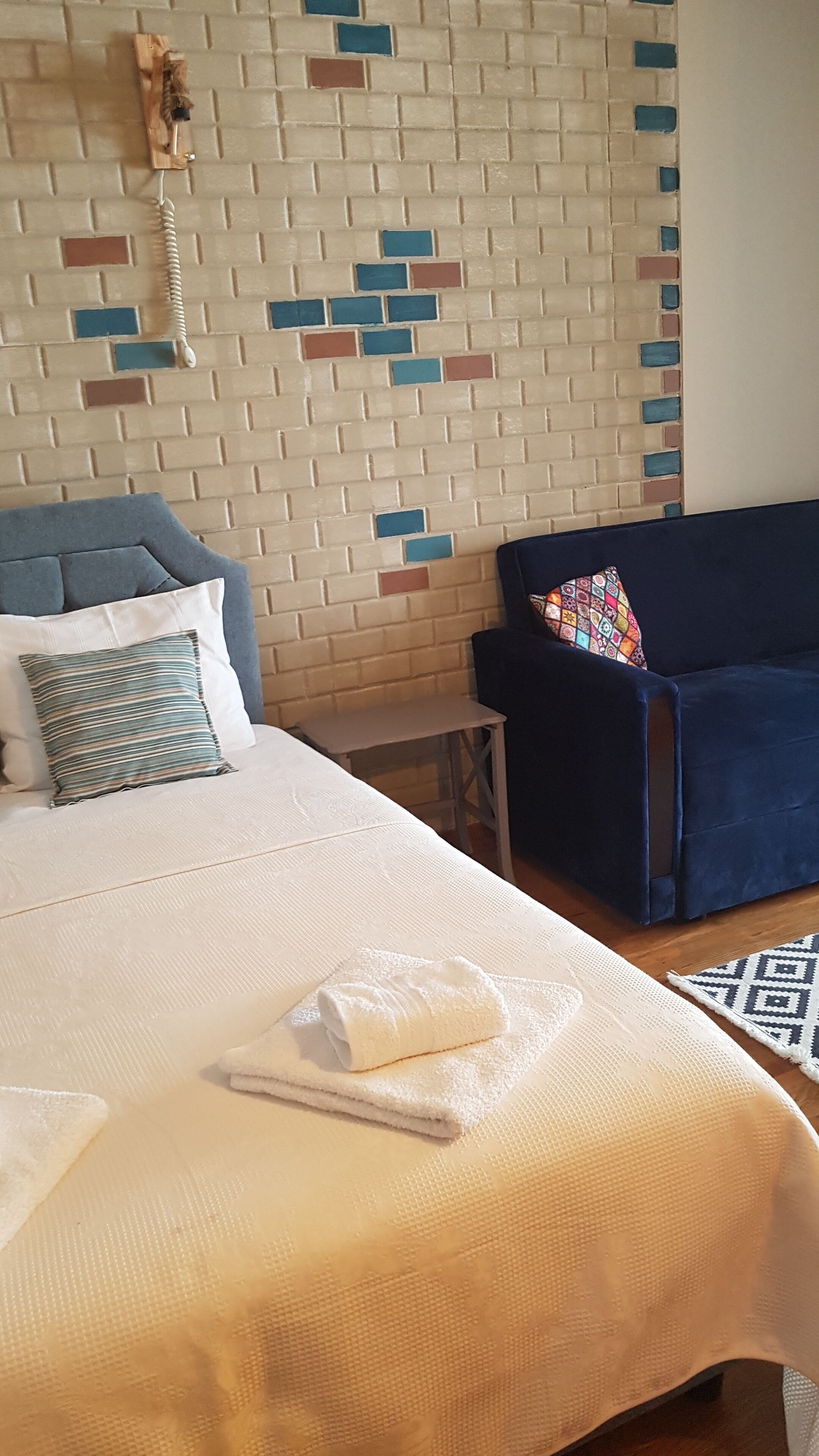 A neatly made bed with a white comforter and a folded white towel. A blue chair with a colorful cushion is positioned against a wall with a patterned tile design.