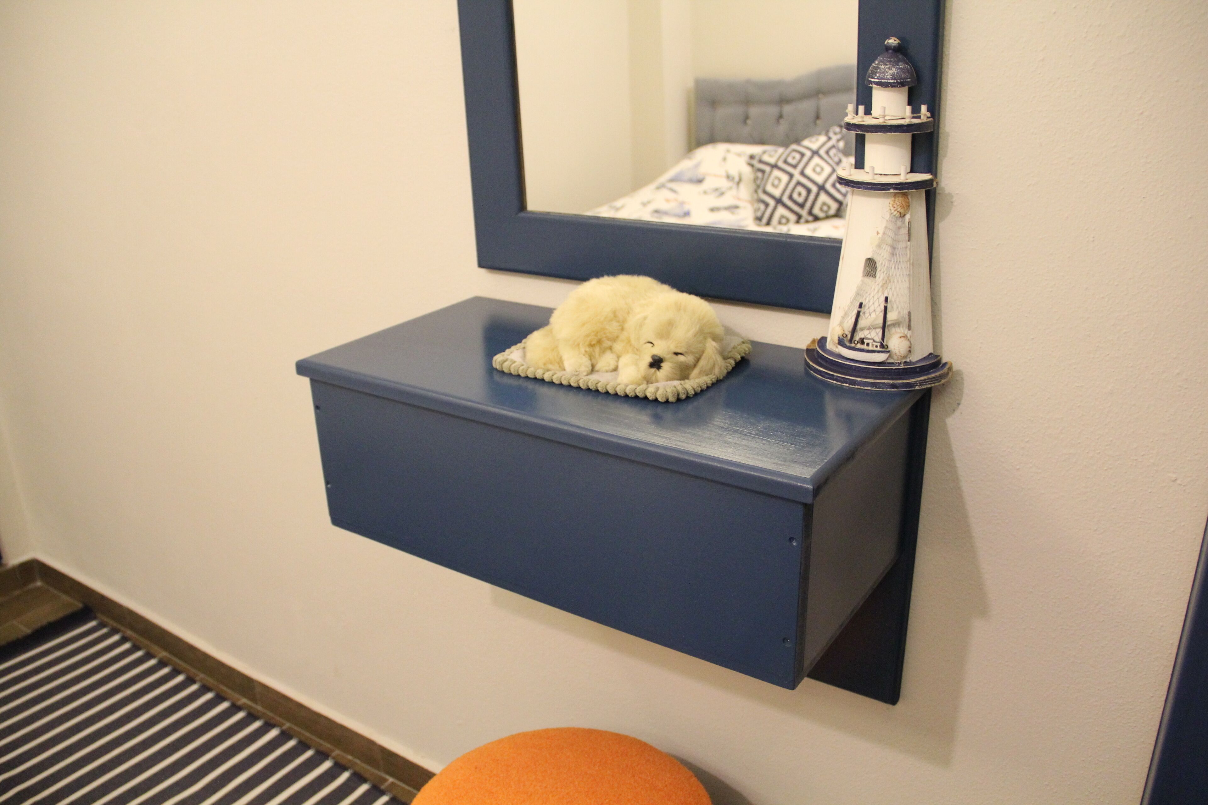 A small white dog resting on a blue shelf with a lighthouse model beside it.
