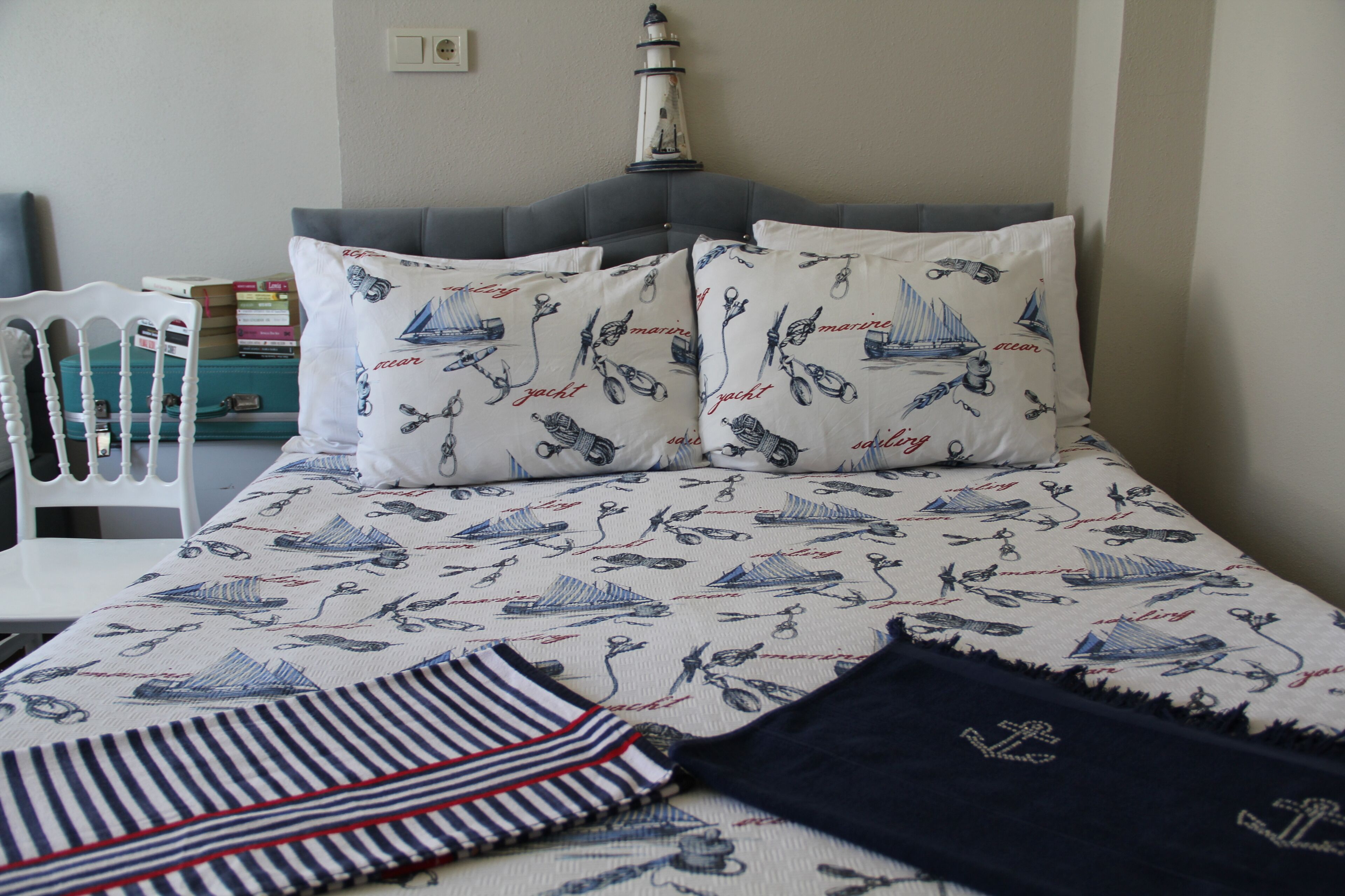 Una cama bien hecha con ropa de cama de temática náutica, una mesita de noche con libros y un faro decorativo en la pared.