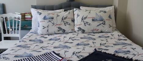 Una cama bien hecha con ropa de cama de temática náutica, una mesita de noche con libros y un faro decorativo en la pared.