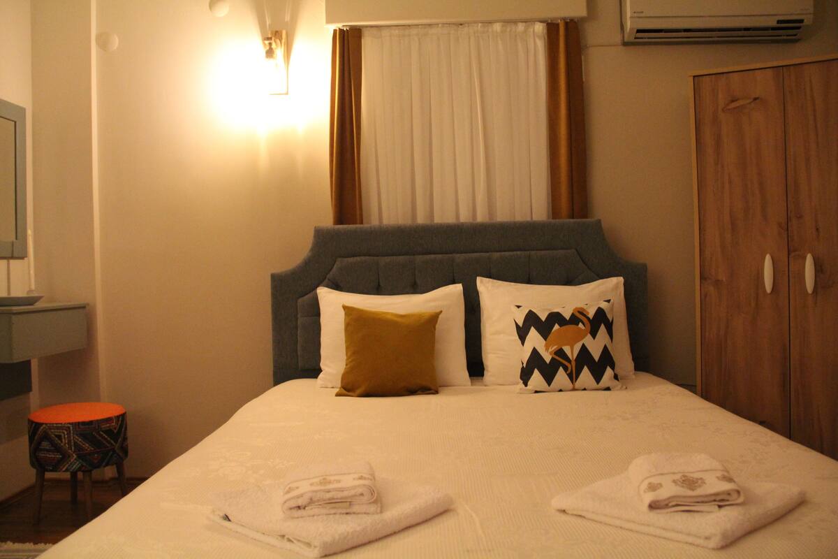 A neatly made bed with a headboard, two pillows, a bedside table, and a small stool.