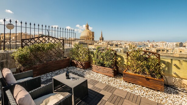 Une terrasse sur le toit, aménagée avec des meubles en osier, des plantes en pot et offrant une vue sur le paysage urbain, notamment un dôme et une flèche imposants.