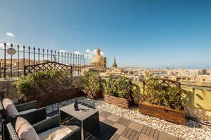 A rooftop terrace with wicker furniture, potted plants, and a view of a cityscape including a prominent dome and spire. - La Vallette (Valletta)